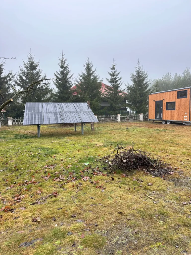 Mobilny domek drewniany na tle lasu, z panelami słonecznymi i stosikiem gałęzi na trawiastym terenie.