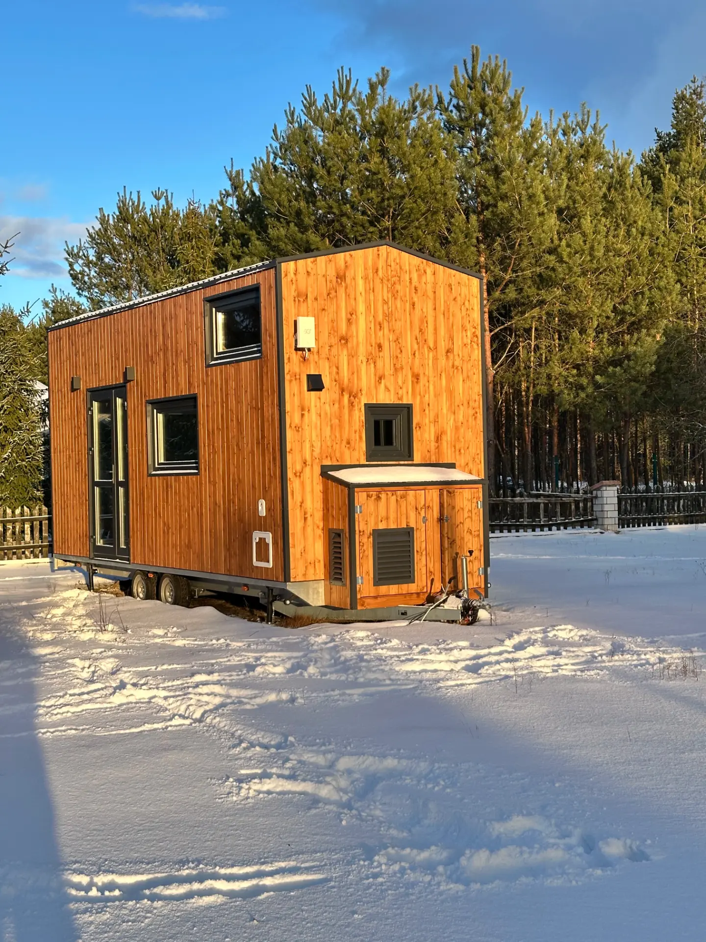 Mobilny domek z drewnianą elewacją na tle lasu iglastego, zaśnieżony teren z widocznymi śladami.