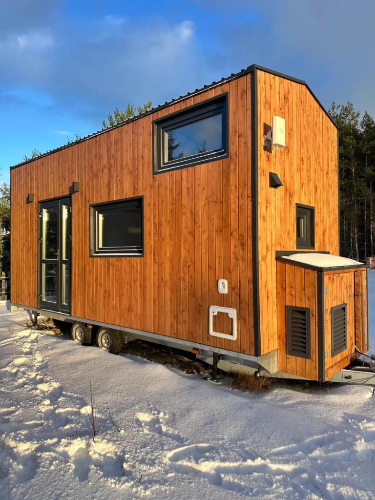 Mobilny domek z drewnianą elewacją, zaśnieżony teren, las w tle i jasne niebo.