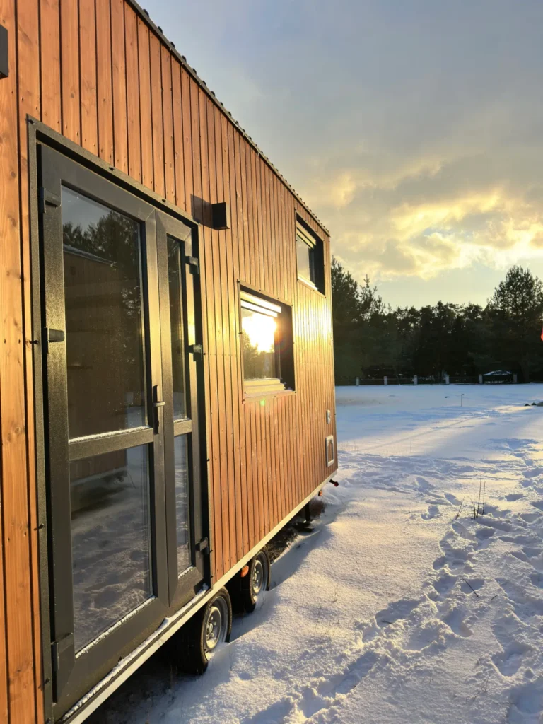 Mobilny domek z drewnianą elewacją na tle zimowego krajobrazu, promienie słoneczne padające na ścianę.