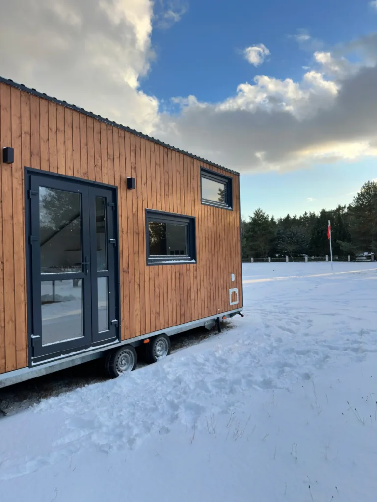 Mobilny domek z drewnianą elewacją na tle lasu, zaśnieżony teren z widoczną flagą.