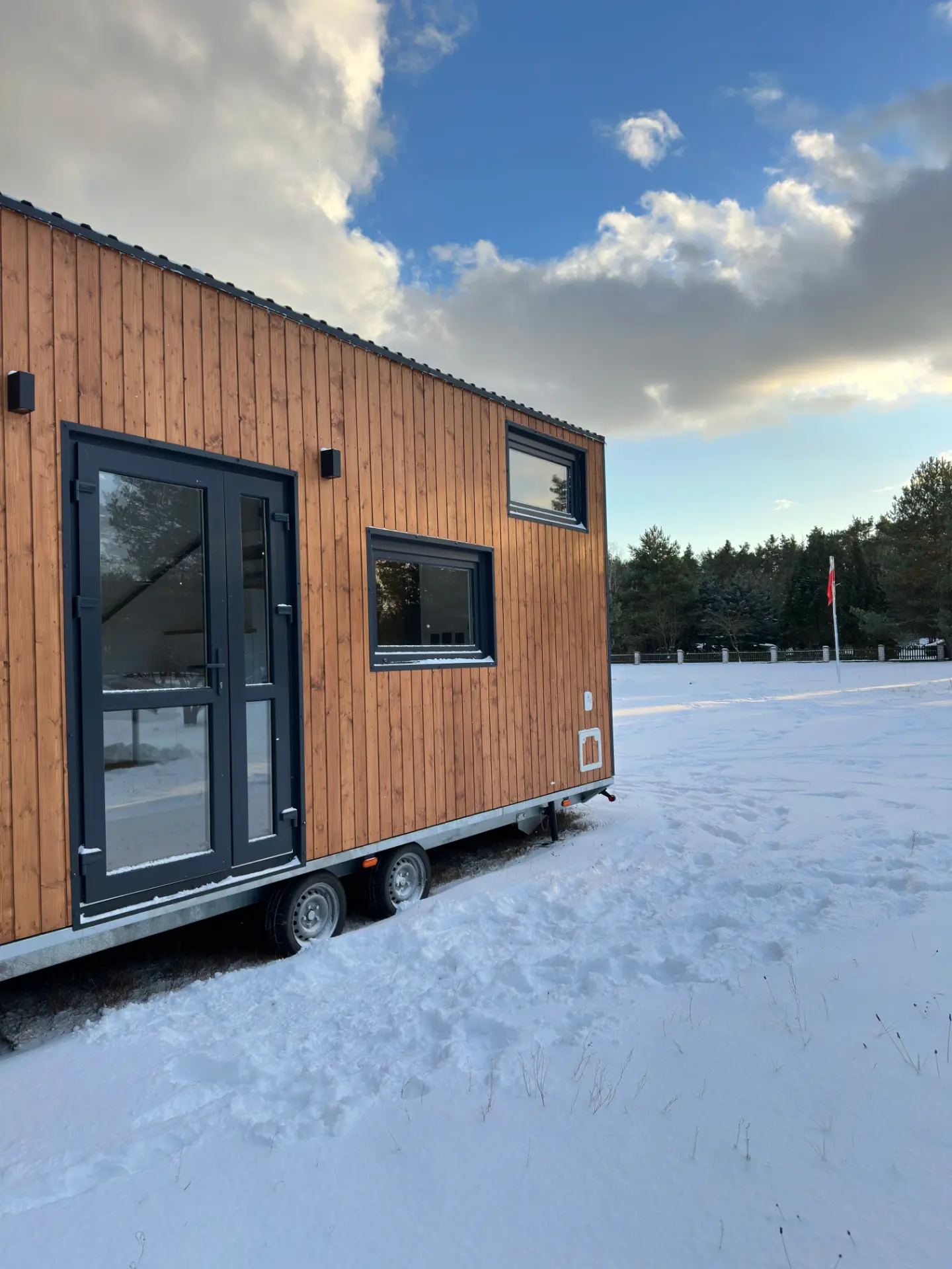 Mobilny domek z drewnianą elewacją na tle lasu, zaśnieżony teren z widoczną flagą.