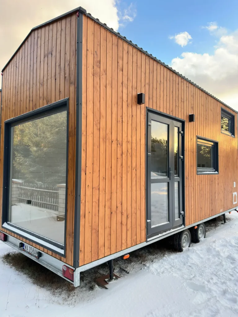 Mobilny domek z drewnianą elewacją, widok na zaśnieżony teren, las i ogrodzenie w tle.